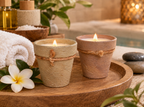 Two lit spa candles in rustic terracotta pots with twine details, styled on a wooden tray with a white towel, plumeria flower, and spa props in a warm bathroom setting.