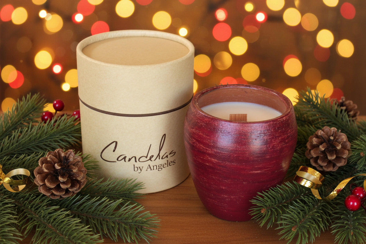 A candle product shown with its kraft paper packaging container and a red ceramic candle vessel on a wooden surface, surrounded by pine branches, pinecones, and festive lights in the background. The container is printed with the brand name 'Candelas by Angeles'.