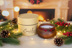 A wooden double-wick candle in a dark brown bowl with red cord trim, shown next to its beige cylindrical cardboard packaging box on a textured beige fabric background. The box is printed with the brand name 'Candelas by Angeles'.