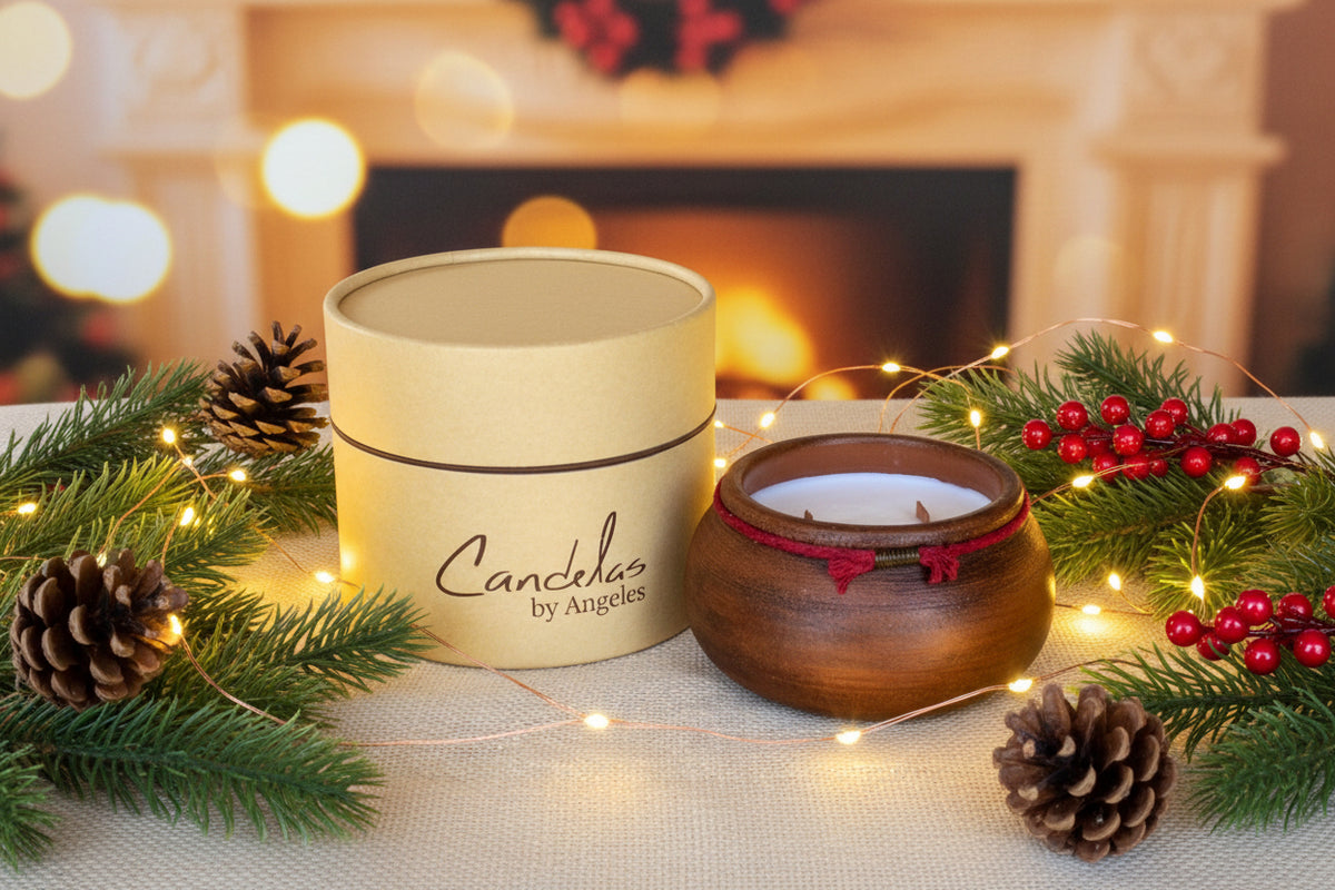 A wooden double-wick candle in a dark brown bowl with red cord trim, shown next to its beige cylindrical cardboard packaging box on a textured beige fabric background. The box is printed with the brand name 'Candelas by Angeles'.