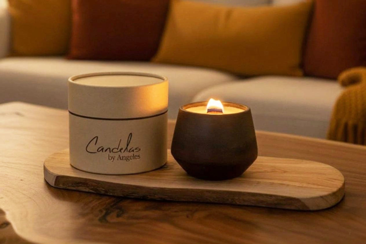A candle product scene showing a white cylindrical candle tin labeled 'Candelas by Angeles' next to a lit dark brown ceramic candle holder on a wooden tray, placed on a wooden table with a blurred living room background.