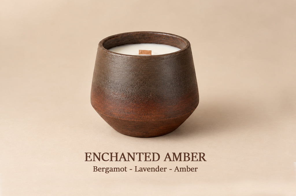 A dark brown ceramic candle vessel with a white wax fill and a small wooden wick, shown on a warm beige background with the text 'ENCHANTED AMBER Bergamot - Lavender - Amber' below.