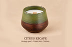 A ceramic candle holder with a green and dark brown glazed finish, containing a white wax candle with a small orange label. The text 'CITRUS ESCAPE' and 'Orange peel - Green tea - Vetiver' are printed below on a beige background.