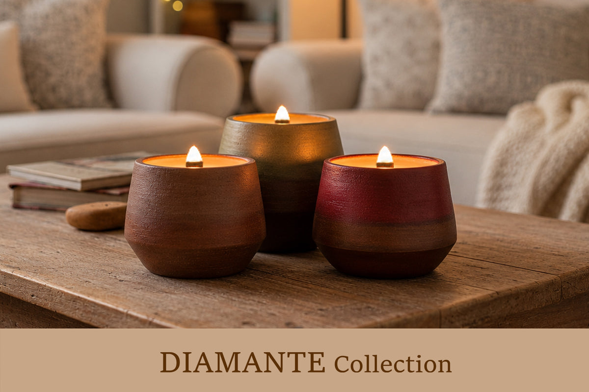 Three lit ceramic candle holders in earthy terracotta, brown, and deep red tones sit on a wooden table in a cozy living room setting. A beige banner at the bottom reads 'DIAMANTE Collection'.