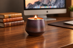 A lit candle in a dark brown textured ceramic holder sits on a wooden desk next to a stack of books, with an iMac and keyboard in the blurred background.