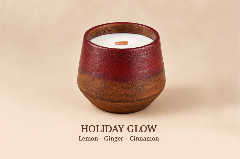A ceramic candle holder with a gradient glaze transitioning from deep red at the top to warm brown at the bottom, containing white wax and a wooden wick. The image is set against a solid beige background with the text 'HOLIDAY GLOW' and 'Lemon - Ginger - Cinnamon' printed below.