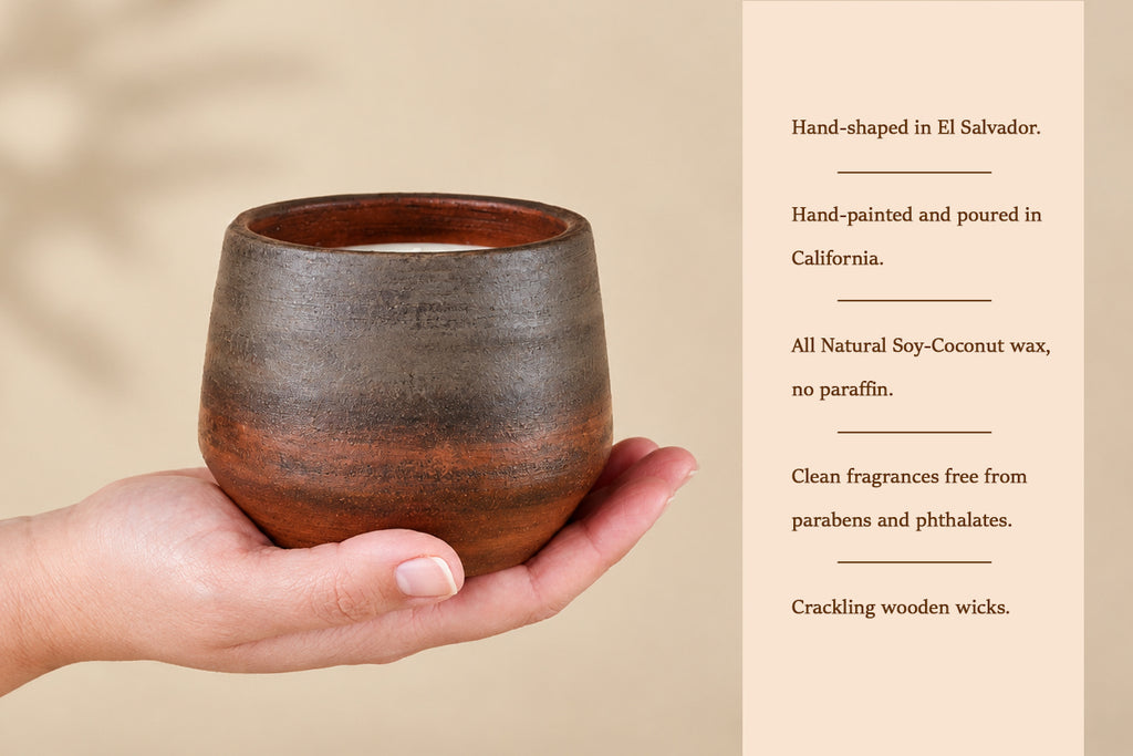 A hand holds a small rustic ceramic candle holder with a brown and black glazed finish, next to a beige text panel describing the candle's hand-shaped El Salvador origin, hand-painted California production, natural soy-coconut wax, and crackling wooden wicks.