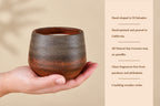A hand holds a small rustic ceramic candle holder with a brown and black glazed finish, next to a beige text panel describing the candle's hand-shaped El Salvador origin, hand-painted California production, natural soy-coconut wax, and crackling wooden wicks.