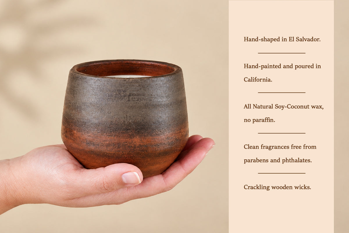A hand holds a small rustic ceramic candle holder with a brown and black glazed finish, next to a beige text panel describing the candle's hand-shaped El Salvador origin, hand-painted California production, natural soy-coconut wax, and crackling wooden wicks.
