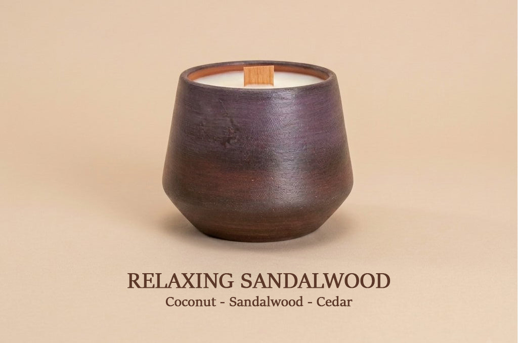 A dark brown ceramic candle vessel with a wooden wick, shown on a warm beige background with the text 'RELAXING SANDALWOOD Coconut - Sandalwood - Cedar' below.