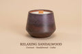A dark brown ceramic candle vessel with a wooden wick, shown on a warm beige background with the text 'RELAXING SANDALWOOD Coconut - Sandalwood - Cedar' below.