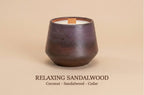 A dark brown ceramic candle vessel with a wooden wick, shown on a warm beige background with the text 'RELAXING SANDALWOOD Coconut - Sandalwood - Cedar' below.