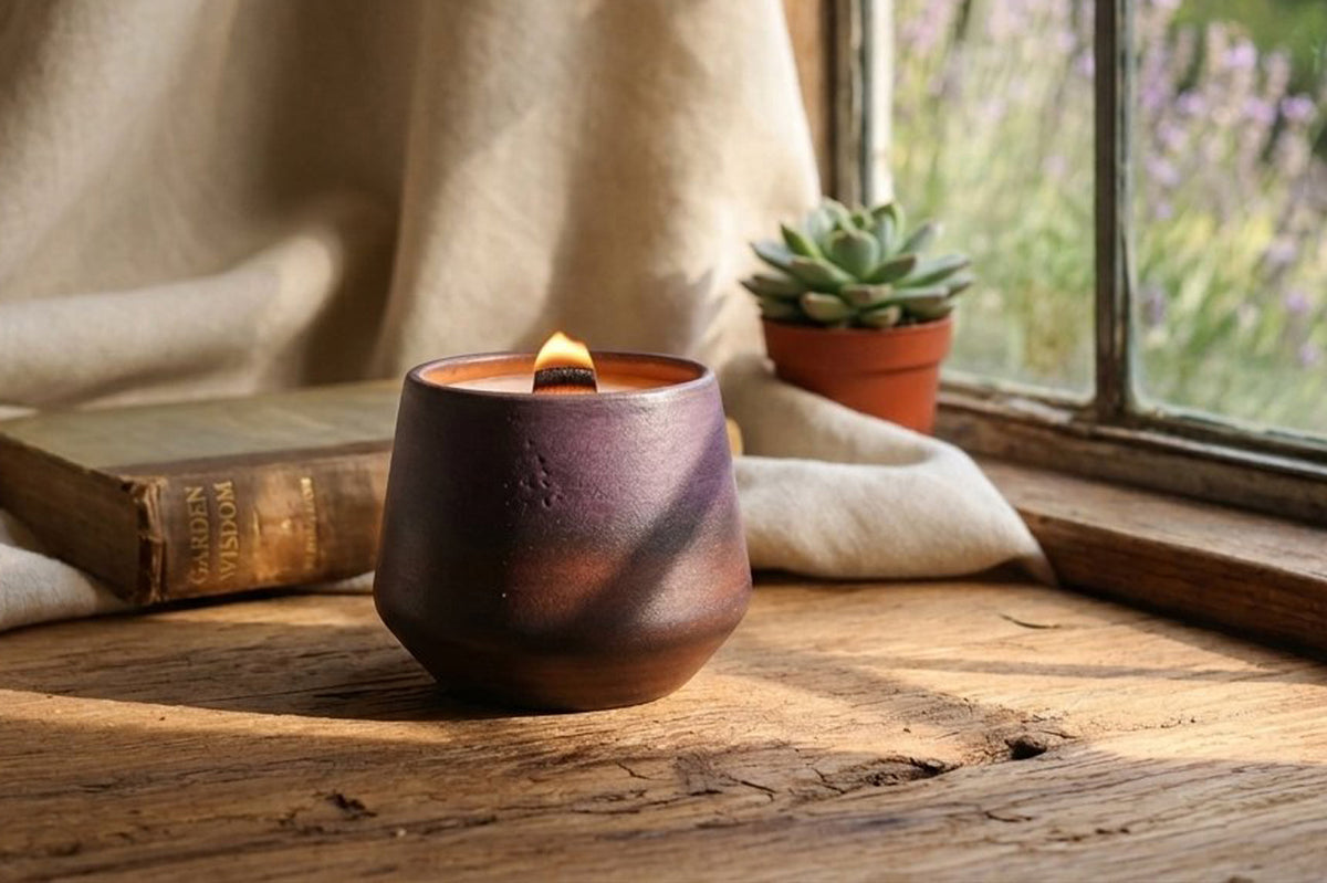 A lit candle in a dark brown textured ceramic holder sits on a rustic wooden table by a window, with a cream linen cloth, an old book, and a small potted succulent in the background.