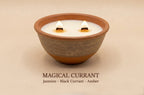 A lit double-wick soy candle in a handmade terracotta bowl with a textured gray-brown middle band, set against a warm beige background. Text below reads 'MAGICAL CURRANT Jasmine - Black Currant - Amber'.