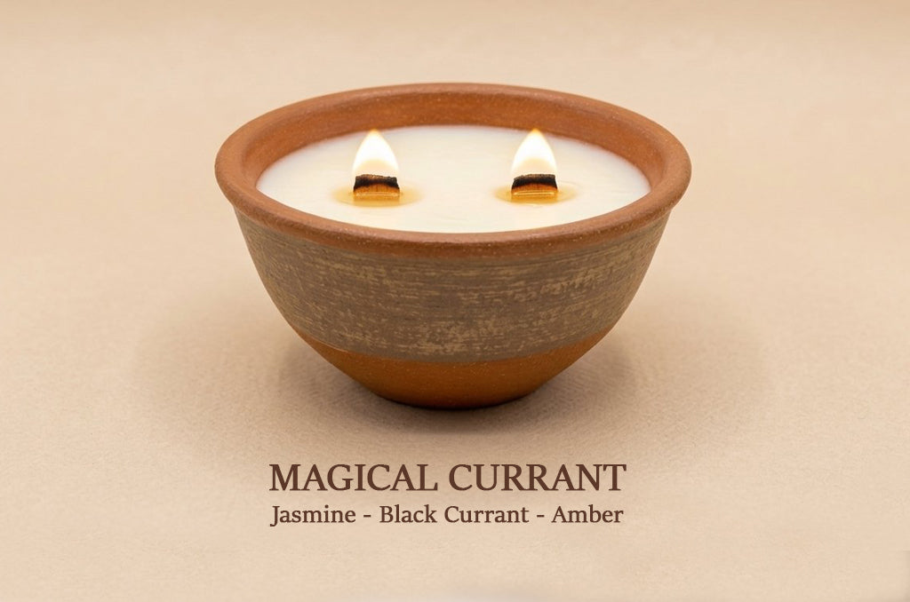A lit double-wick soy candle in a handmade terracotta bowl with a textured gray-brown middle band, set against a warm beige background. Text below reads 'MAGICAL CURRANT Jasmine - Black Currant - Amber'.