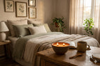 A cozy bedroom scene with a neatly made bed in soft beige and sage green bedding, framed botanical prints on the wall, a bedside lamp, and a wooden table in the foreground holding a lit candle bowl, an open book, a mug, and a spoon. Sunlight streams in from a window with sheer curtains and potted plants.