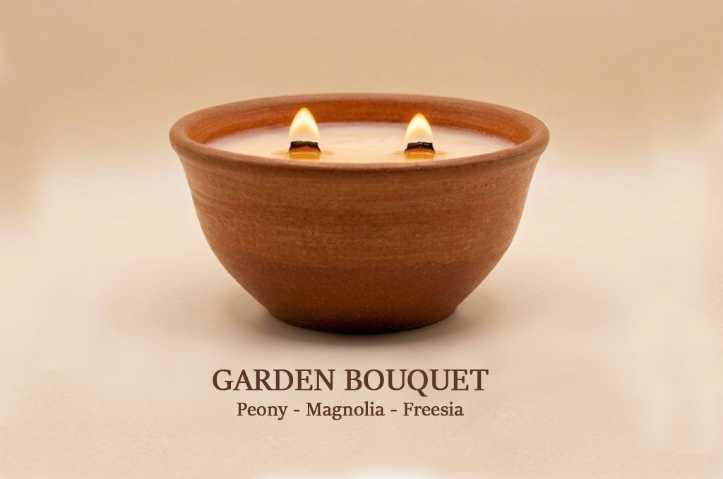 A terracotta-colored ceramic bowl candle with two lit wicks, shown against a warm beige background with the text 'GARDEN BOUQUET Peony - Magnolia - Freesia' below.