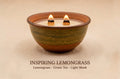 A ceramic bowl candle with two lit wicks, shown on a warm beige background with the text 'INSPIRING LEMONGRASS Lemongrass - Green Tea - Light Musk' below.