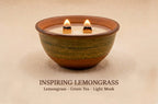 A ceramic bowl candle with two lit wicks, shown on a warm beige background with the text 'INSPIRING LEMONGRASS Lemongrass - Green Tea - Light Musk' below.
