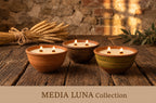 Three lit wax candles in rustic ceramic bowls arranged on a wooden table, with wheat and greenery in the background and a banner reading MEDIA LUNA Collection at the bottom.