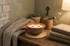 A lit two-wick candle in a textured terracotta bowl sits on a woven coaster on a rustic wooden table, surrounded by a knitted throw blanket, books, a mug, and a small potted succulent in a cozy room setting.