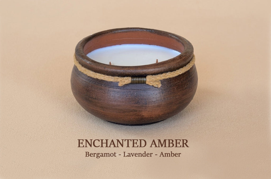 A dark brown wooden candle holder with a white wax candle inside, featuring a natural jute rope accent tied in a bow. The image is set against a solid beige background with the text 'ENCHANTED AMBER' and 'Bergamot - Lavender - Amber' printed below.