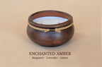 A dark brown wooden candle holder with a white wax candle inside, featuring a natural jute rope accent tied in a bow. The image is set against a solid beige background with the text 'ENCHANTED AMBER' and 'Bergamot - Lavender - Amber' printed below.