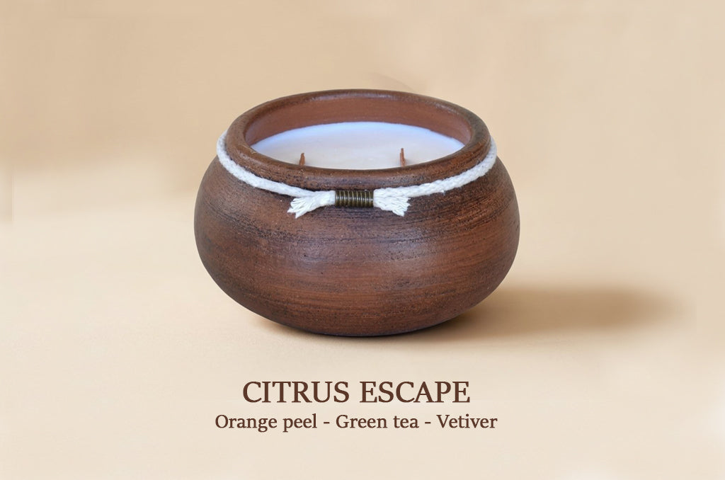 A round dark brown wooden candle holder with a white cord and small bead detail, containing a white wax candle. The image is set against a warm beige background with the text 'CITRUS ESCAPE Orange peel - Green tea - Vetiver' below.