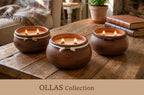 Three rustic brown clay candles in round pots with white rope accents, lit and displayed on a wooden table in a cozy living room setting. A banner at the bottom reads 'OLLAS Collection'.