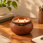 A lit two-wick candle in a round, rustic terracotta-colored ceramic pot with a dark textured band and small metal accent, set on a wooden desk with a plant and stationery in the background.