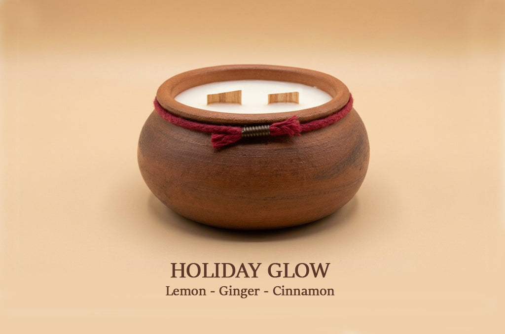 A small round wooden holder or candle vessel with a white interior and two visible wicks, tied with a dark red cord, on a warm beige background. Text below reads 'HOLIDAY GLOW' and 'Lemon - Ginger - Cinnamon'.