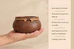A hand holds a small round brown wooden vessel with a rope-wrapped rim on a beige background, next to a text panel describing the product's handcrafted El Salvador origin, California pouring, natural soy-coconut wax, and crackling wooden wicks.