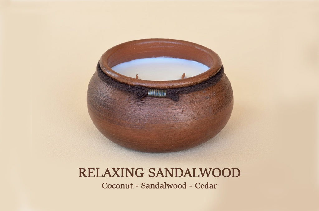 A round wooden candle holder or vessel with a warm brown sandalwood finish, a dark band around the upper portion, and a pale coconut-colored wax pool inside. Text below reads 'RELAXING SANDALWOOD Coconut - Sandalwood - Cedar'.
