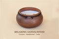 A round wooden candle holder or vessel with a warm brown sandalwood finish, a dark band around the upper portion, and a pale coconut-colored wax pool inside. Text below reads 'RELAXING SANDALWOOD Coconut - Sandalwood - Cedar'.