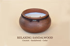 A round wooden candle holder or vessel with a warm brown sandalwood finish, a dark band around the upper portion, and a pale coconut-colored wax pool inside. Text below reads 'RELAXING SANDALWOOD Coconut - Sandalwood - Cedar'.