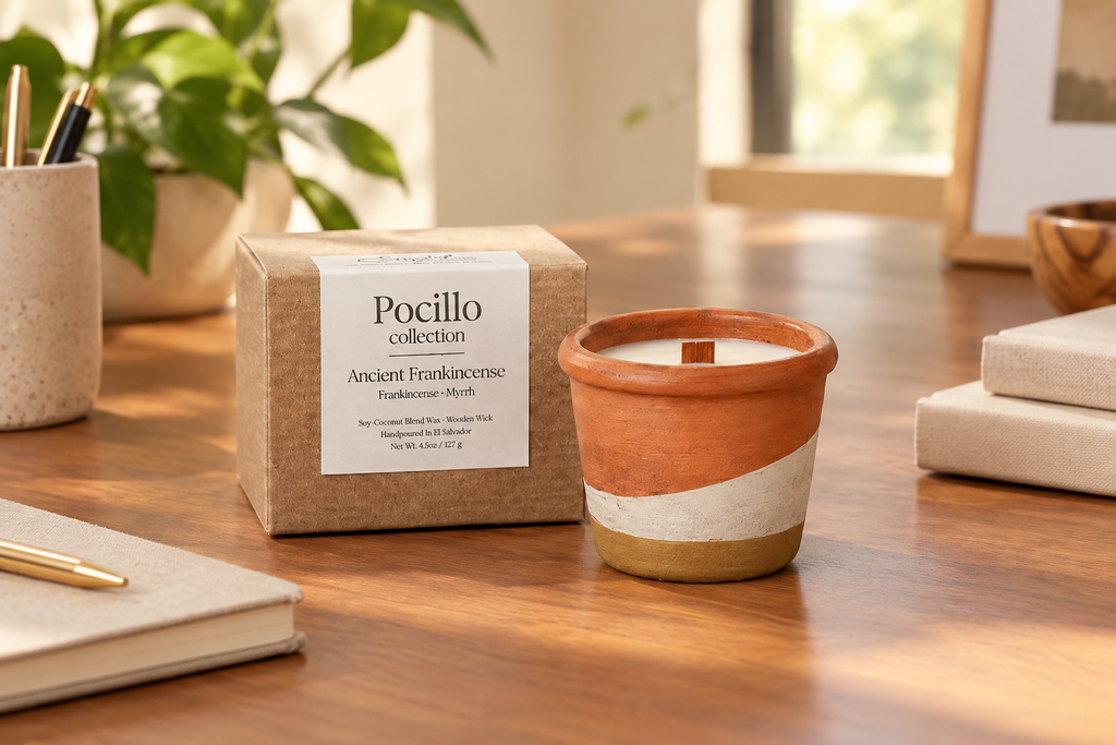 A Pocillo Collection candle in a terracotta and cream ceramic pot sits on a wooden desk next to its kraft paper box packaging, with a blurred office background.