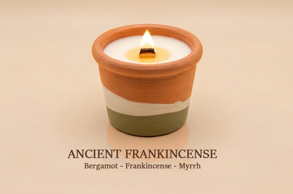 A lit candle in a terracotta-colored ceramic holder with a white wax top and a white band, set against a warm beige background. Text below reads 'ANCIENT FRANKINCENSE' with 'Bergamot - Frankincense - Myrrh'.