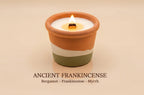 A lit candle in a terracotta-colored ceramic holder with a white wax top and a white band, set against a warm beige background. Text below reads 'ANCIENT FRANKINCENSE' with 'Bergamot - Frankincense - Myrrh'.
