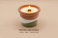 A lit ceramic candle in a terracotta-colored pot with a white wax top and a white and sage green band around the middle, shown on a beige background with the text 'FRESH ORCHID Citrus - Mint - Tomato Leaf' underneath.