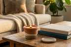 A lit terracotta candle holder with a small flame sits on a wooden coffee table in a cozy living room setting, next to stacked books and a small dish, with a sofa and potted plant in the background.