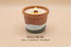 A lit scented candle in a terracotta-colored ceramic pot with a white wax top and a dark gray lower band, shown on a warm beige background with the text 'WILD MUSK Citrus - Sandalwood - Dark Musk' below.