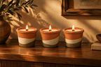Three lit candles in terracotta-colored ceramic holders with horizontal beige and gray bands, arranged on a wooden table in a warmly lit interior setting.