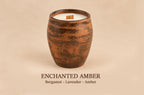 A single candle in a rustic amber-brown ceramic vessel with white wax and a small wooden wick, centered on a warm beige background. Text below reads 'ENCHANTED AMBER' and 'Bergamot - Lavender - Amber'.