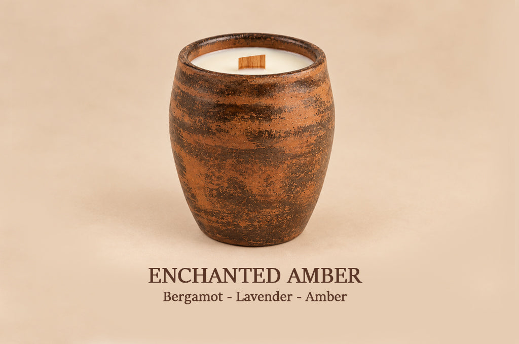 A single candle in a rustic amber-brown ceramic vessel with white wax and a small wooden wick, centered on a warm beige background. Text below reads 'ENCHANTED AMBER' and 'Bergamot - Lavender - Amber'.
