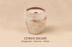A ceramic jar candle with a textured beige and brown glaze, filled with white wax and featuring a small orange wood wick. The text 'CITRUS ESCAPE' and 'Orange peel - Green tea - Vetiver' is printed below on a solid beige background.