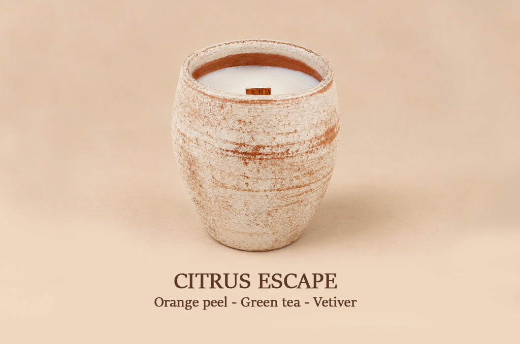 A ceramic jar candle with a textured beige and brown glaze, filled with white wax and featuring a small orange wood wick. The text 'CITRUS ESCAPE' and 'Orange peel - Green tea - Vetiver' is printed below on a solid beige background.