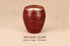 A wooden candle holder with a textured reddish-brown finish, containing white wax and a small wooden wick, set against a solid beige background with the text 'HOLIDAY GLOW Lemon - Ginger - Cinnamon' below.