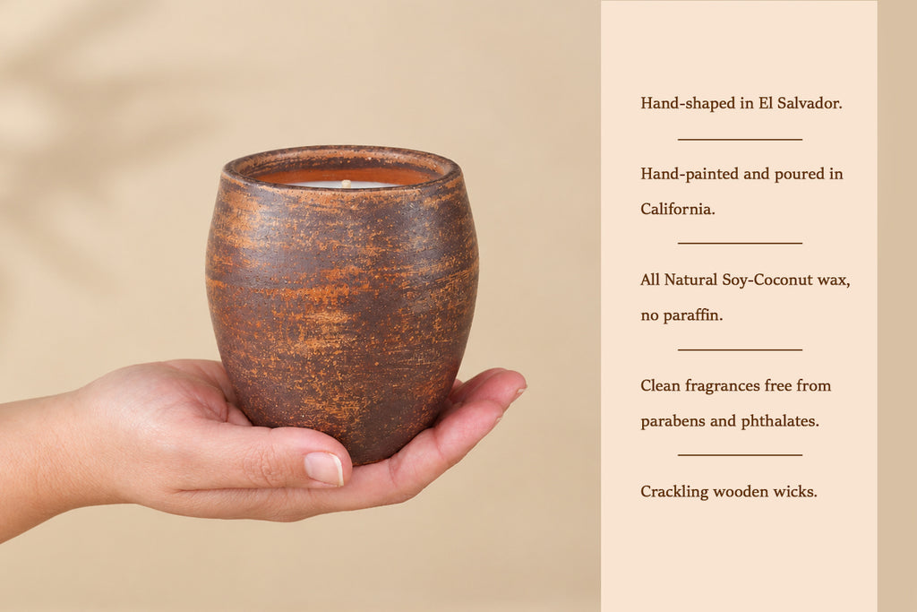 A hand holds a small round brown ceramic candle holder with a textured finish, next to a beige text panel describing the candle's hand-shaped El Salvador origin, hand-painted California pouring, soy-coconut wax, and crackling wooden wicks.