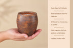 A hand holds a small round brown ceramic candle holder with a textured finish, next to a beige text panel describing the candle's hand-shaped El Salvador origin, hand-painted California pouring, soy-coconut wax, and crackling wooden wicks.