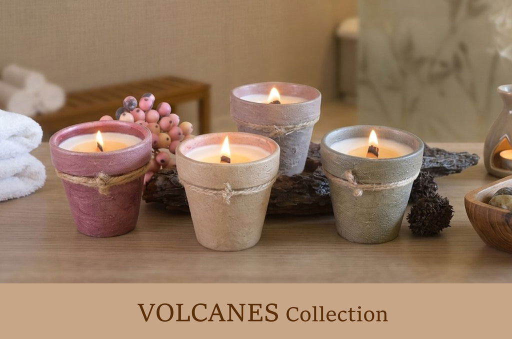 Four lit wax candles in rustic volcano-shaped holders on a wooden surface, with a spa-like bathroom setting in the background and the text 'VOLCANES Collection' at the bottom.
